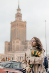 Fototapeta premium A young girl in glasses with flowers against the background of the Palace of Culture in Warsaw. A pretty girl in glasses with flowers walks in Warsaw.