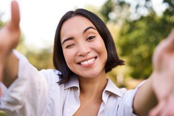 People and mobile connection. Happy young asian woman takes selfie on smartphone, holds camera with hands, poses in park on summer day