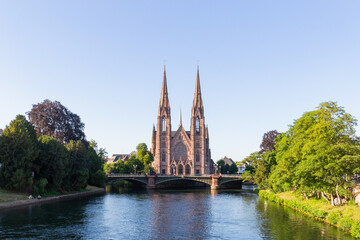 Church with two towers in France surrounded by a river, built with a reddish stone and near trees