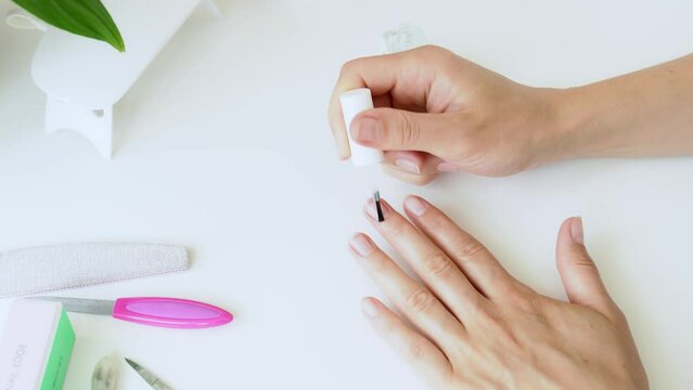 Girl applying colorless nail polish to her nails close up - home beauty treatment. The young woman covering female nails with colorless varnish for protection before use UV lamp. Wellness spa concept