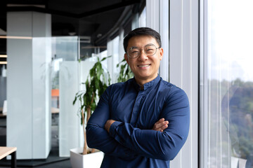 Portrait of successful asian businessman, man inside office wearing shirt and glasses smiling and looking at camera, investor broker with crossed arms near window.