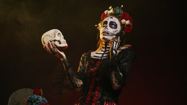 Mexican horror goddess on phone call holding skull, talking on smartphone line and acting creepy in studio. Answering telephone for remote chat, wearing holy mexican entity costume. Handheld shot.