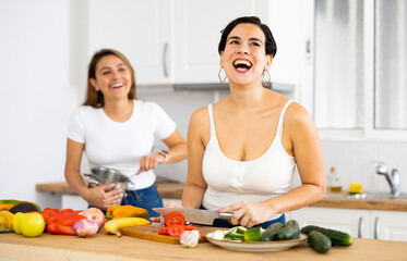 Obraz na płótnie Canvas Smiling young Hispanic woman cooking with sister in home kitchen, preparing fresh vegetable salad. Happy family relationship concept