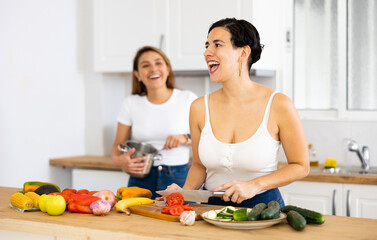 Obraz na płótnie Canvas Happy young latin american girl standing in cozy kitchen interior at home with girlfriend, slicing fresh vegetables for salad for dinner. LGBT couple relationship concept..