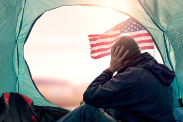 Homeless man in dark clothes sitting inside a tent USA flag and town out of focus in the background. Hands on his head in despair. Social issue. Poverty concept. Sad refugee in hard living conditions.