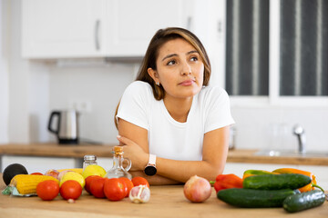 Portrait of upset frustrated young Hispanic woman standing in home kitchen, leaning on table full of fresh vegetables