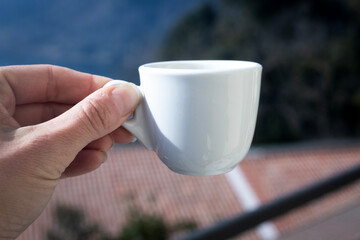 Human hand with small white empty cup espresso coffee in bright sunlight. Sky and orange roof in background out of focus. Cups up. Caffeine energy drink. Meditation moment. Coffee brake. 