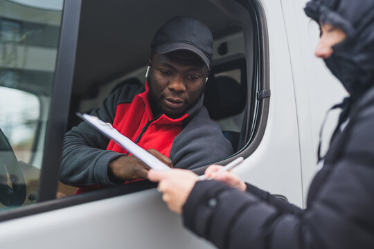 Person in puffy jacket signing papers held out the window of white van by black young adult delivery man wearing red pullover. Horizontal shot . High quality photo