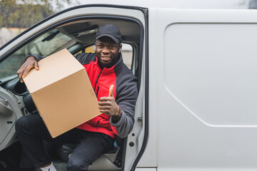 Young adult black delivery man getting out of white van with carboard box parcel looking into camera smiling and showing thumbs up. Horizontal shot. High quality photo