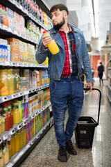 Young glad positive smiling bearded man choosing bottled fresh juices in grocery section at supermarket