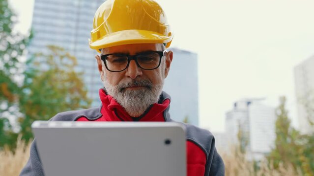 Caucasian, bearded, grey-haired professional construction worker in uniform wearing a bright orange helmet and eyeglasses, standing, holding a tablet used for work. High quality 4k footage