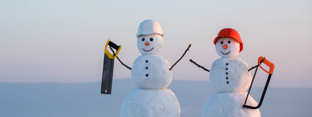 Two snowmen builders in building helmet hold saw. Snowman in hard hat on the snow outdoor...
