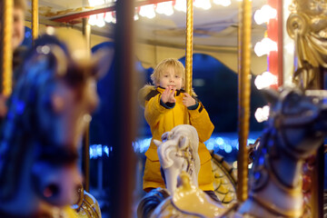 Cute blonde boy enjoying Christmas fair. Little child riding on a vintage carousel (merry go...
