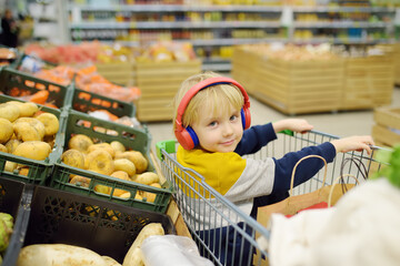 Cute preschooler boy with headphone and player is sitting in a shopping cart at a food store or supermarket. A child is listening to music or an audiobook while his parents buying groceries.