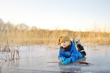 Cute preschool boy is playing on the ice of a frozen lake or river on a cold sunny winter sunset. Child having fun with icicle and dry reed plant during family hiking. Kids outdoor games - obrazy, fototapety, plakaty