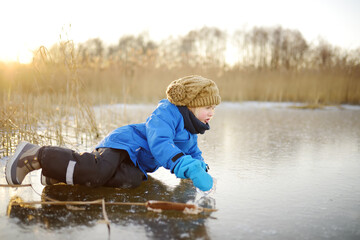 A cute preschooler fell on the ice of a frozen lake or river at a cold sunny winter sunset