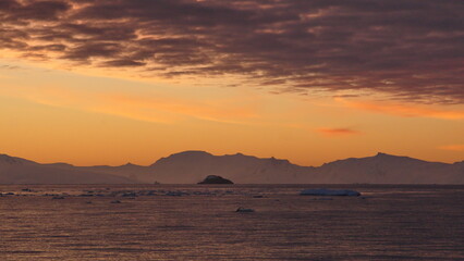 Naklejka na ściany i meble Pink clouds in an orange sky, with mountains and icebergs in silhouette, at Cierva Cove, Antarctica, at sunset