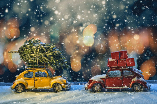 Two models of old passenger cars with a Christmas tree and gifts on the roof stand under the snow in the evening electric light.
