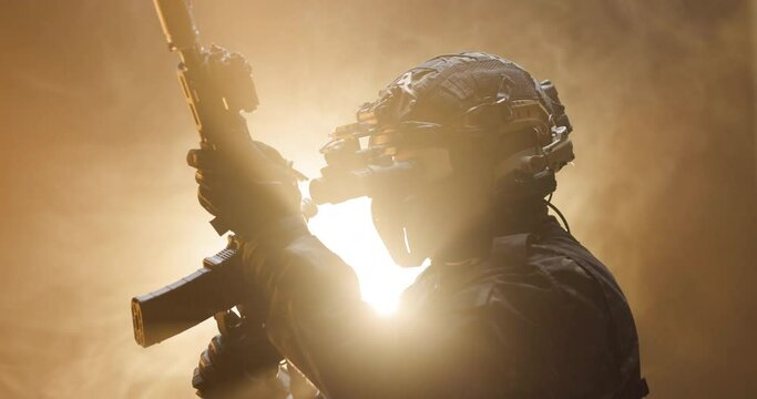 Soldier fully equipped with tactical gear and protection aiming his rifle at enemy with orange smoke in the background.