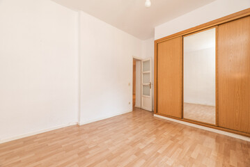 Empty room with wooden built-in wardrobe with cherry-colored sliding doors and white wooden access door and matching laminated flooring