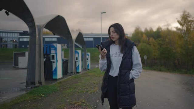 Woman at charging station. Charging her EV by using her phone