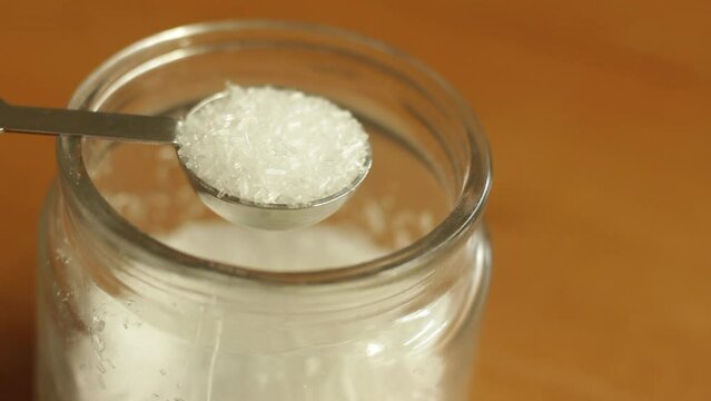 A teaspoon of white refined MSG monosodium glutamate powder is being scooped with stainless steel measuring spoon cup