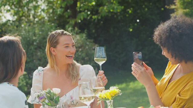 Woman taking photo on mobile phone of friends sitting in summer garden drinking wine and celebrating with a cheers  whilst chatting and catching up - shot in slow motion