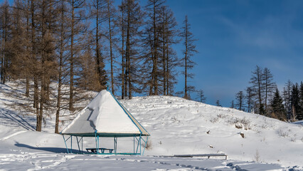 In the valley there is a gazebo with a layer of snow on a pyramidal roof. Bare trees on a hill against a blue sky. Altai