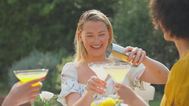Three female friends sitting outdoors in summer garden at home making a toast with cocktails - shot in slow motion 
