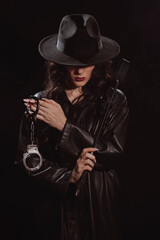 Fototapeta premium Sexy female dominant mistress with whip flogger and handcuffs for BDSM sex with submission and domination in a leather raincoat and hat in noir style