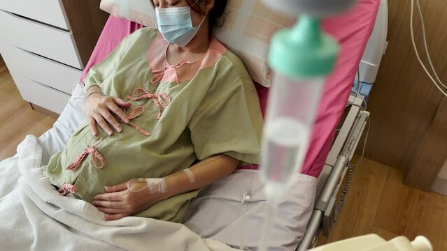 pregnant woman with medical mask stroking her belly on a bed in hospital, protection covid-19 (coronavirus concept)