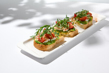 Aesthetic composition with shrimp bruschetta on white background with shadows from flowers. Italian bruschetta with shrimp, tomatoes and arugula on fine dining in summer. Elegant menu concept.