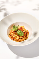 Vegetarian pasta with basil and tomatoes on white plate. Tomato pasta in summer menu with hard shadows Spaghetti with tomatoes on light background. Vegan food - spaghetti marinara
