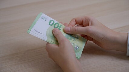 Women's hands with a neat manicure slowly count paper money with a denomination of one hundred euros over the table, then combine the bills into one pile