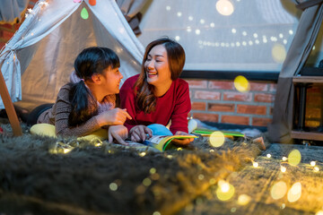Asian mother and cute smiling girl reading a book together while lying in illuminated tent in kid bedroom. Cheerful ethnic woman and lovely daughter playing under a cozy hut.