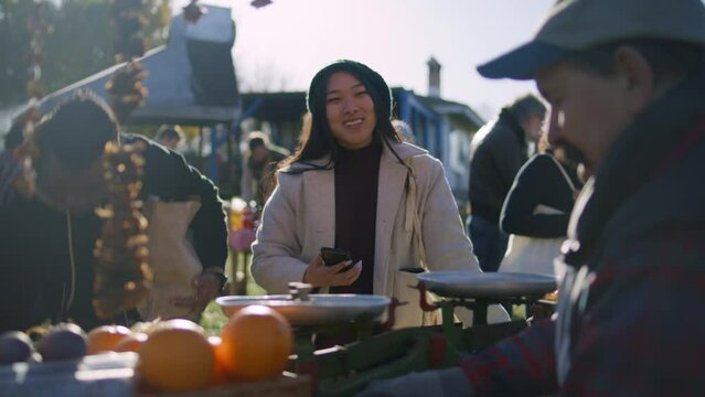 Senior farmer gives paper bag with goods to African American man. Asian woman contactless pays for purchases using smartphone. Shopping at local farmers market on weekend. Vegetarian and organic food.