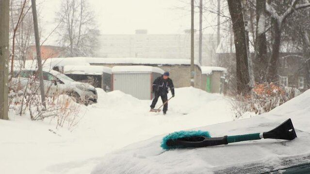 A man in defocus clears the snow with a shovel, it is snowing. There is a car brush. Everything is covered with snow after a snowstorm, snowdrifts all around. City life, heavy snowfall. UHD 4K.