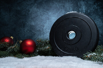 Dumbbell weight plate, red Christmas baubles and tree branches on white snow. Fitness holiday...