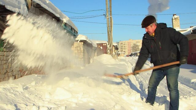 A man deftly clears the path to the garage from snow with a wooden shovel. Large snowdrifts around after a snowstorm, sunny day. City life, heavy snowfall. UHD 4K.