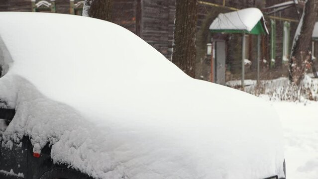 A man with a neat brush cleans the hood of a car from snow, it is snowing. The car is covered with snow after a snowstorm and looks like a snowdrift. City life, heavy snowfall. UHD 4K.