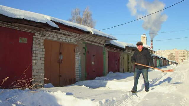 A man clears the path to the garage from snow with a wooden shovel. Large snowdrifts around after a snowstorm, sunny day. City life, heavy snowfall. UHD 4K.