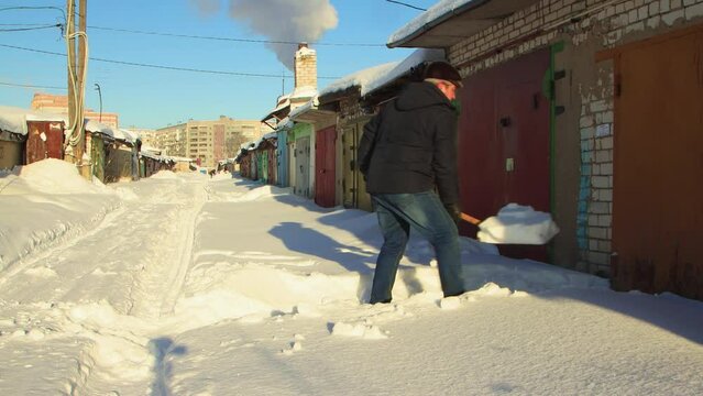 A cheerful man clears the passage to the garage from snow with a wooden shovel. Large snowdrifts around after a snowstorm, sunny day. City life, heavy snowfall. UHD 4K.