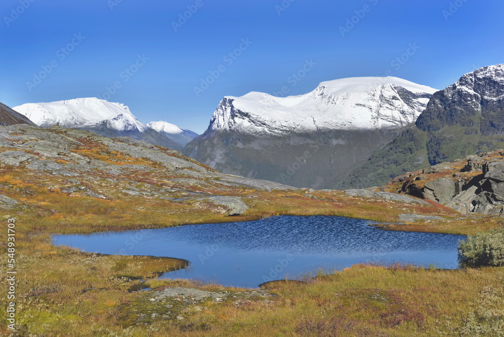 Wall mural small blue lake nestled in the snow-capped norwegian mountains and surrounded by autum-coloured vege