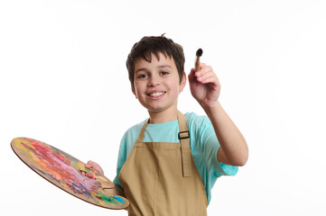 Artistic teenage boy wearing beige apron, holding a paintbrush and a color palette, paints on...