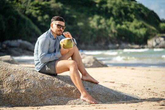 Healthy food in the morning..A man in glasses drinking fresh coconut milk on the beach in the morning barefoot sitting on a stone and looking at the camera
