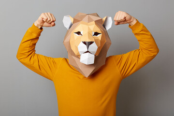 Indoor shot of strong confident man with lion mask with wearing casual style orange jumper, standing with raised arms, showing his power, posing isolated over gray background.