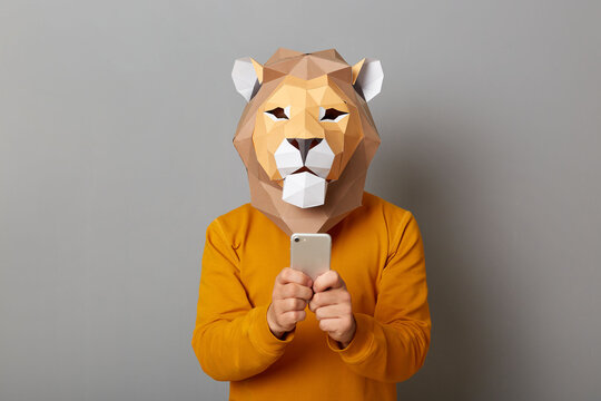Image of man with lion mask with wearing orange sweater, standing with cell phone in hands, using mobile phone for browsing Internet or chatting in social networks posing isolated over gray background