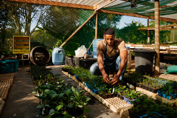 Young black man crouching next to plants while gardening in garden center