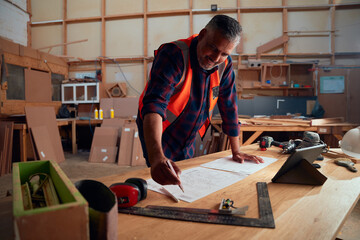 Mid adult man writing on paper between tools and timber in woodworking factory