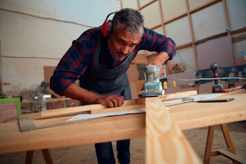Mid adult man wearing ear muffs using power tool on timber plank in woodworking factory